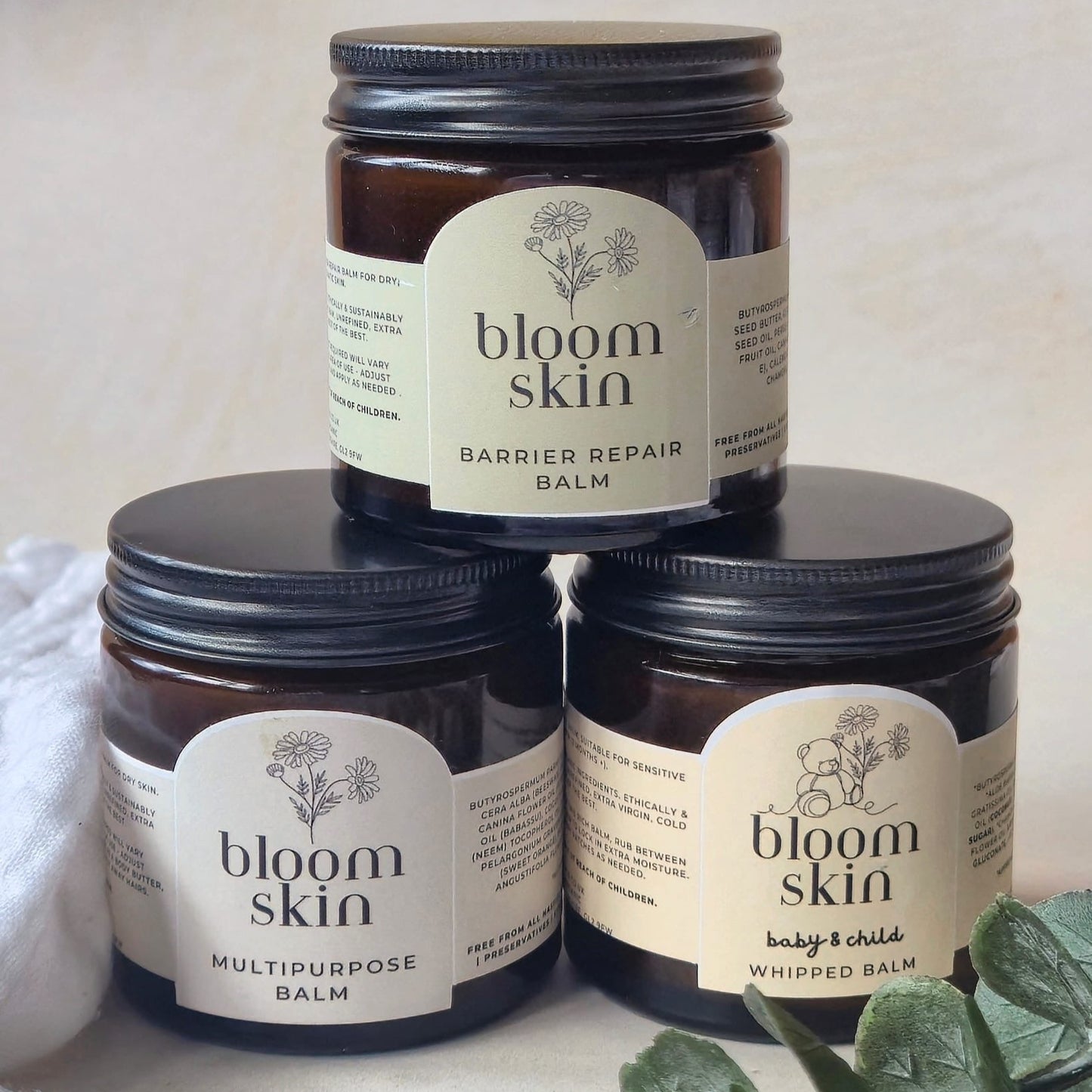Three jars of 'bloom skin' balm stacked on a light background with a plant.