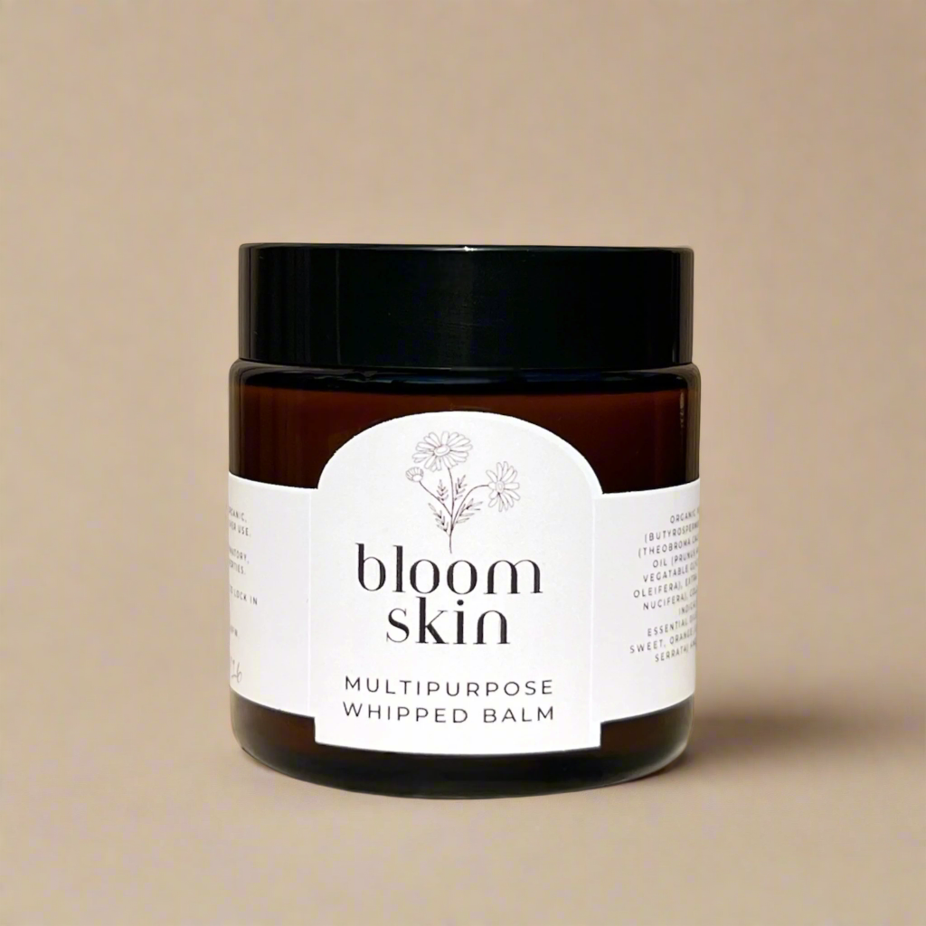 Jar of bloom skin whipped balm on a wooden surface with flowers and an orange slice in the background.