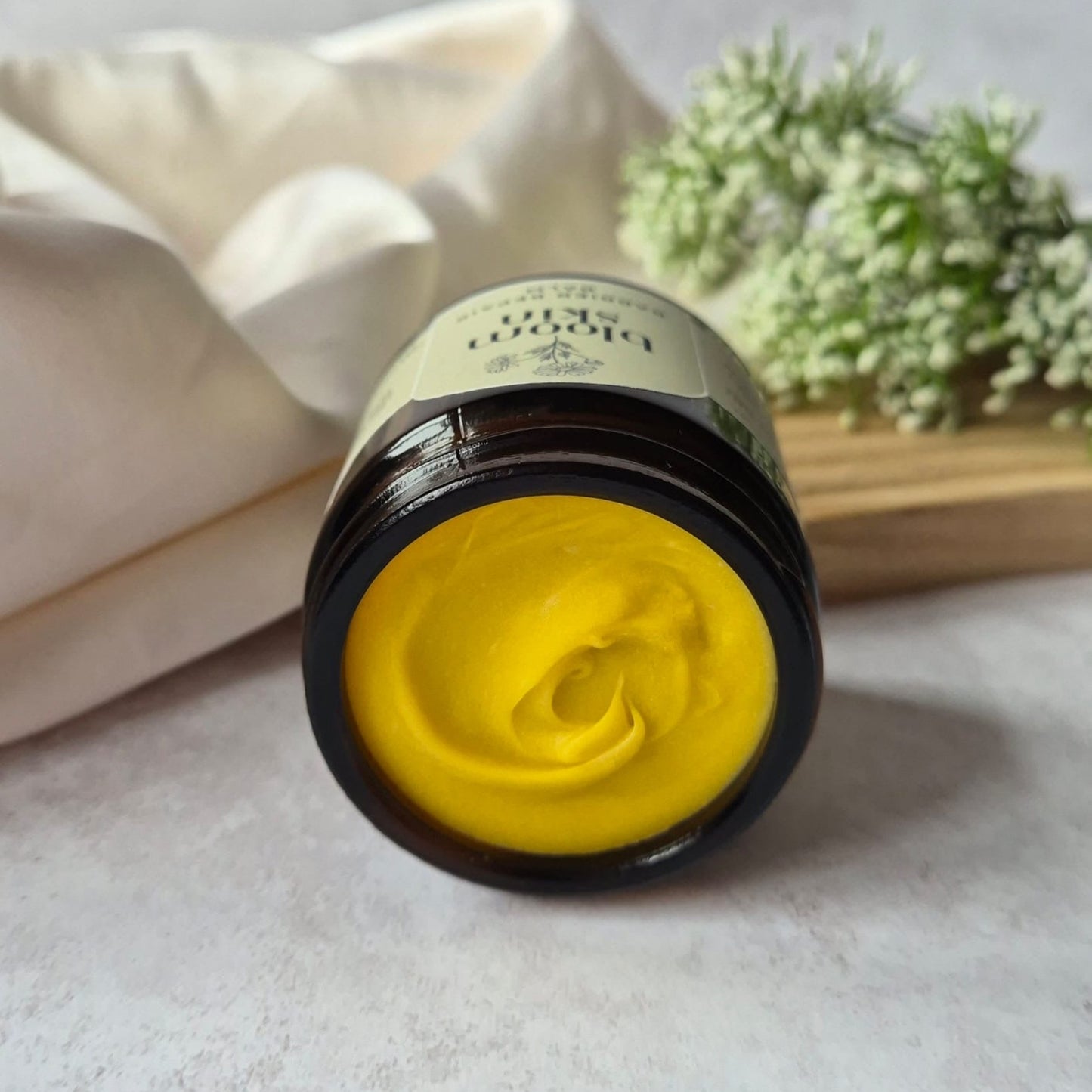 Jar of yellow cream with a black lid on a light surface, with a white cloth and greenery in the background.