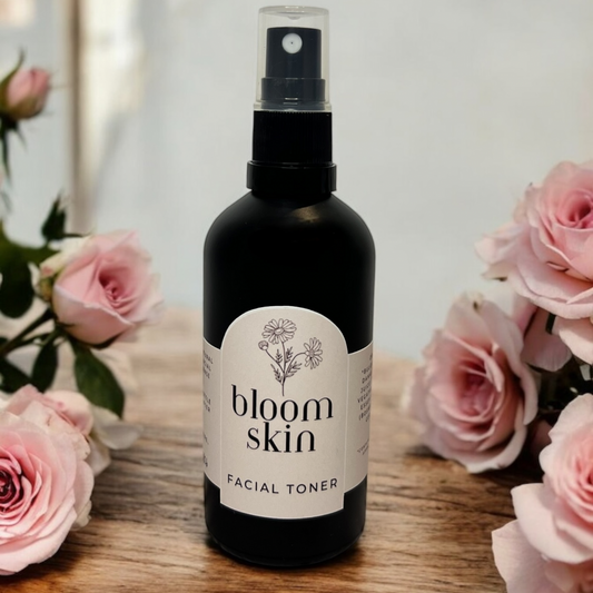 A black glass bottle of Bloom Skin Facial Toner with a spray dispenser, labeled with white and pink text, placed on a wooden surface with pink roses in the background.