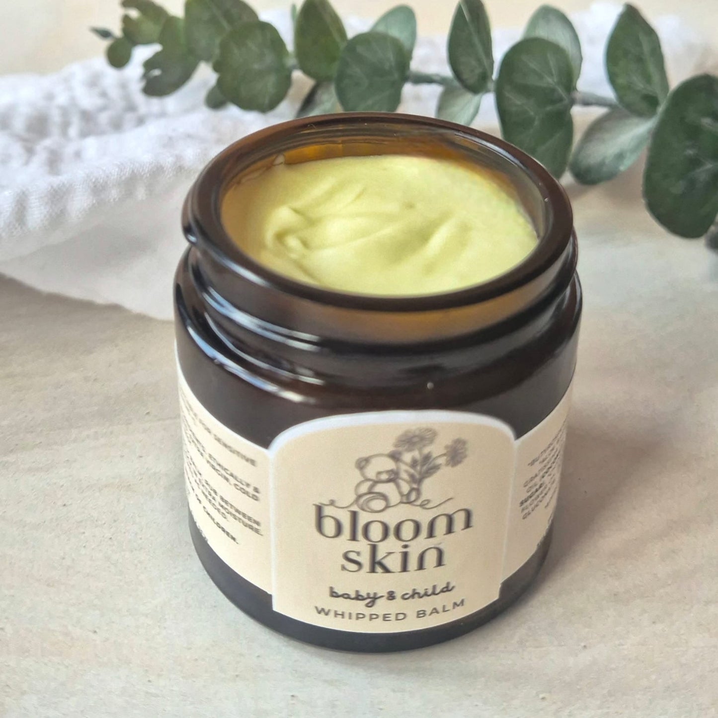 Jar of 'bloom skin' whipped balm on a light surface with eucalyptus leaves in the background.