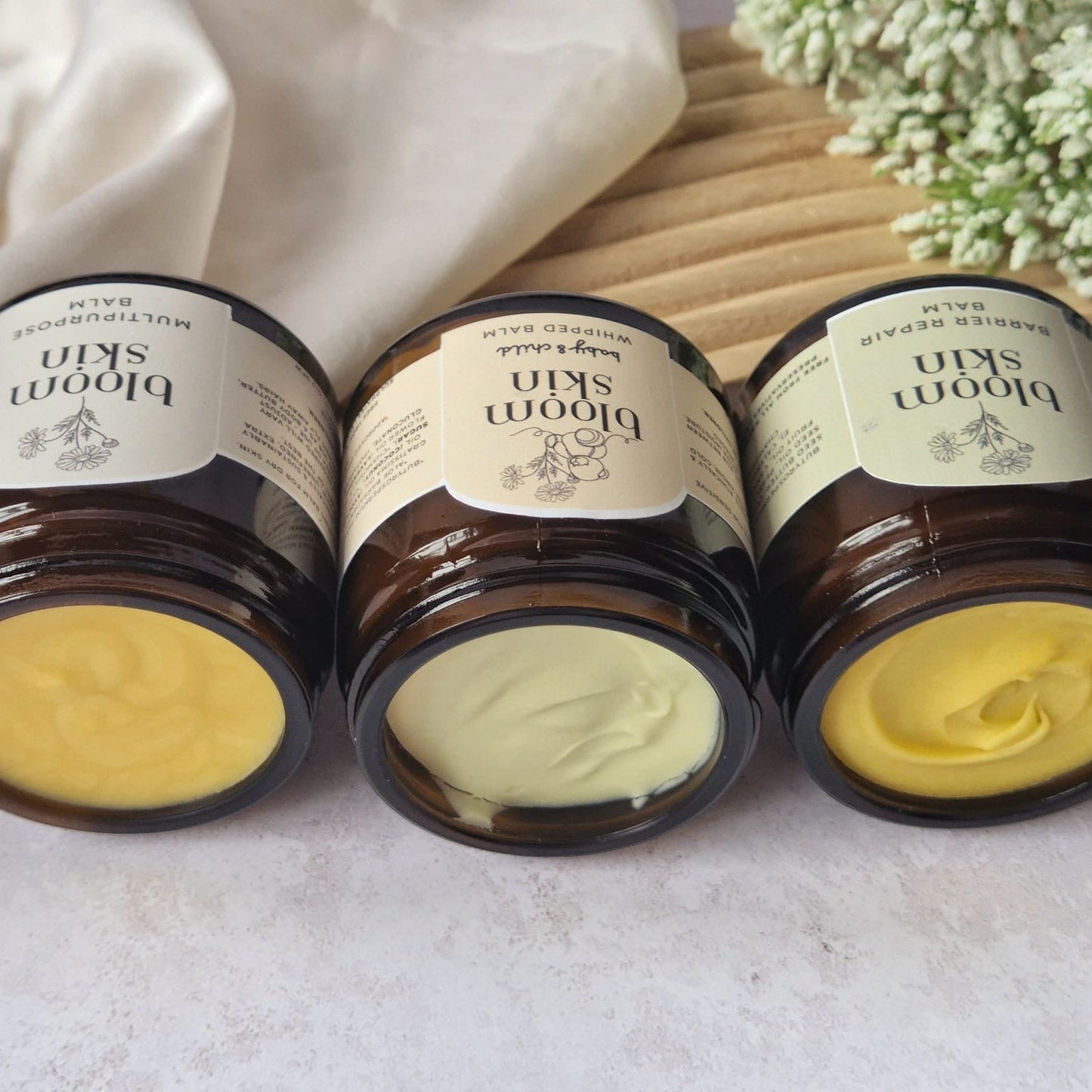 Three jars of Broom Skin cream on a light background with flowers and fabric.