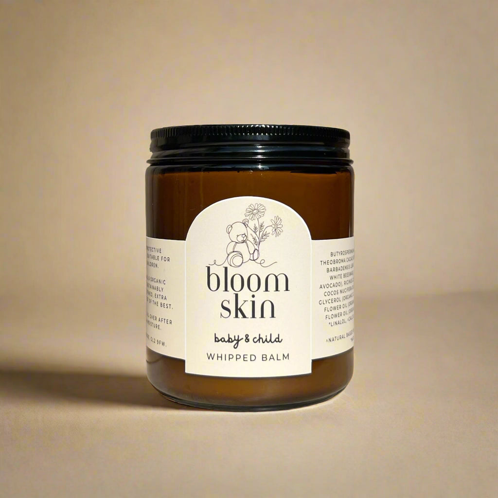 Jar of whipped balm labeled 'bloom skin' on a wooden surface with flowers in the background.