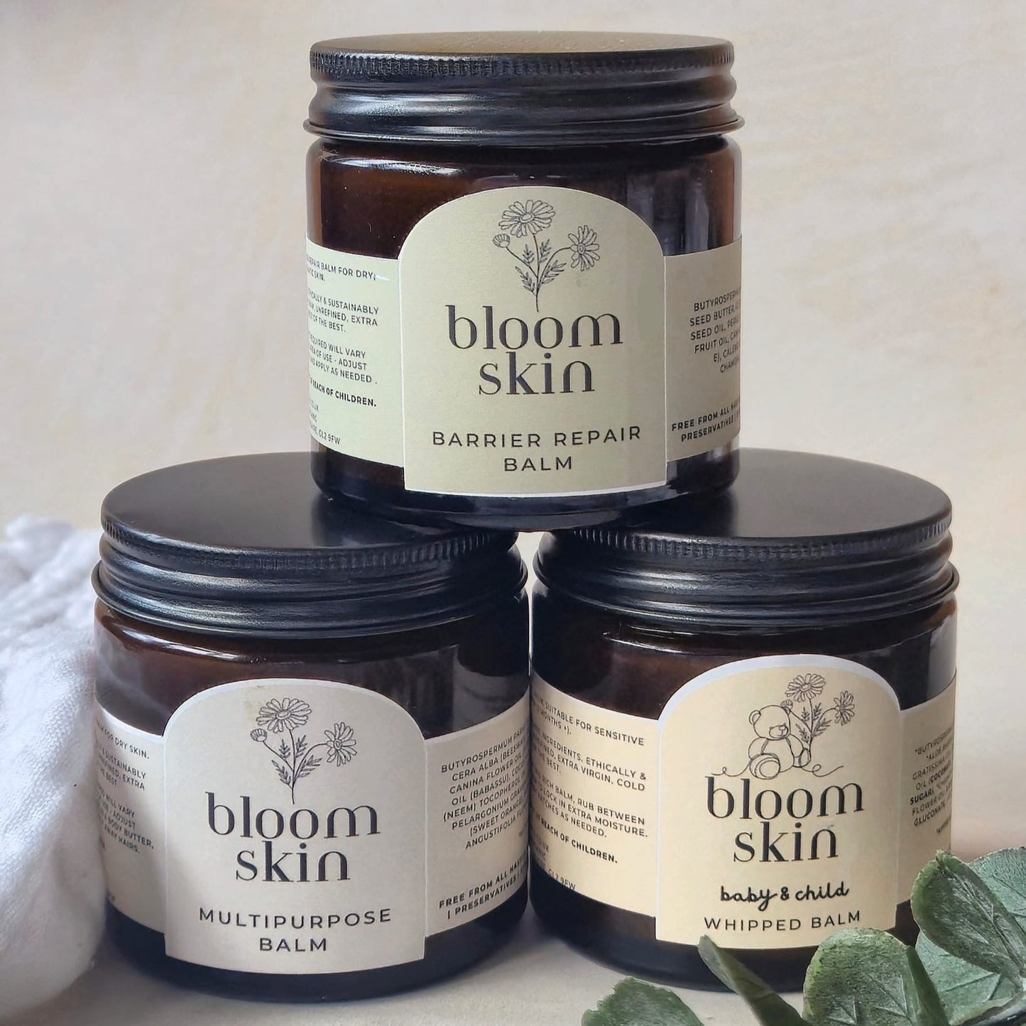 Three jars of 'bloom skin' balm stacked on a light background with greenery.