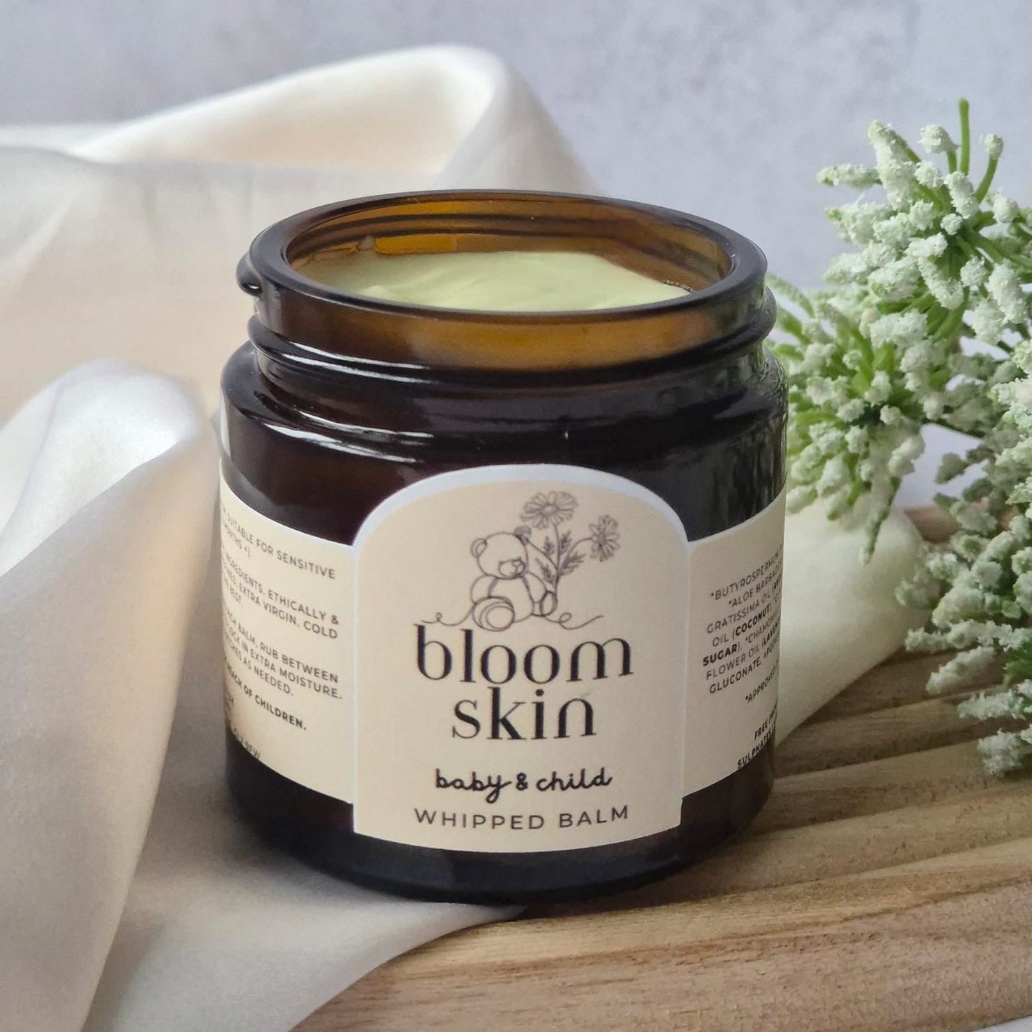 Jar of 'bloom skin' whipped balm on a wooden board with a white cloth and flowers in the background.