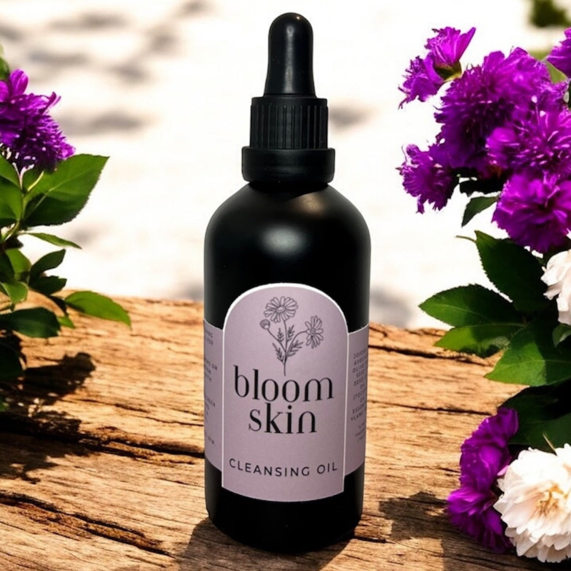 A bottle of Bloom Skin Cleansing Oil placed on a wooden surface with a background of purple and white flowers.