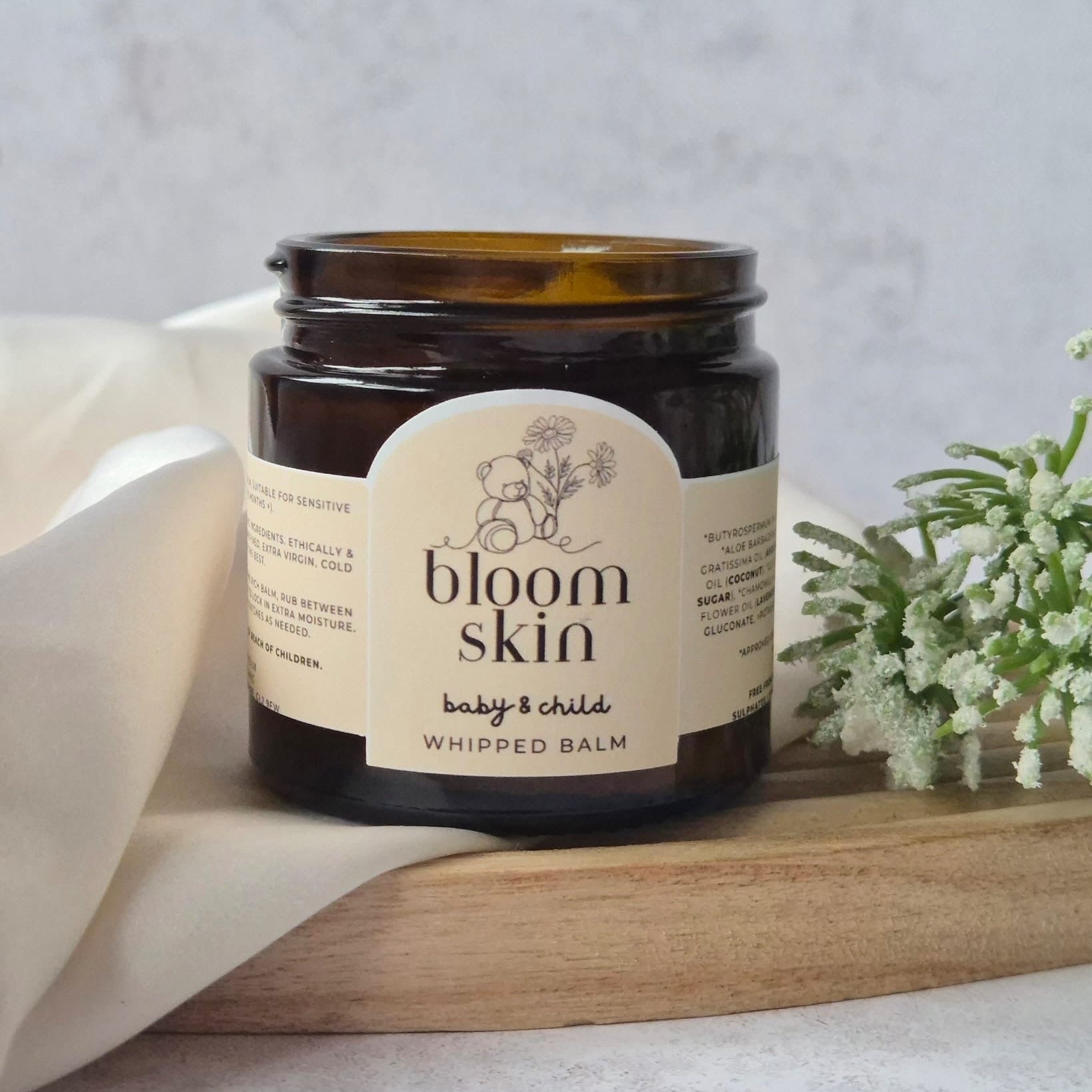 Jar of 'bloom skin' baby and child whipped balm on a wooden board with a white cloth and flowers in the background.