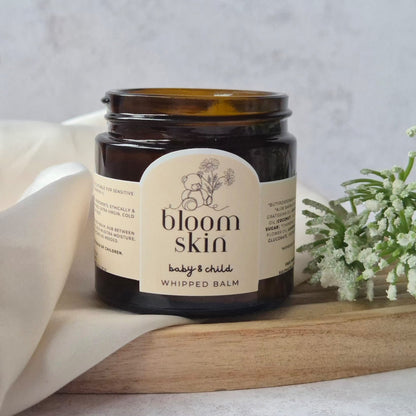 Jar of 'bloom skin' baby and child whipped balm on a wooden board with a white cloth and flowers in the background.